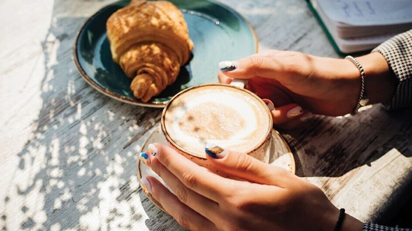 En person holder i en kopp kaffe ved siden av en croissant på en tallerken.