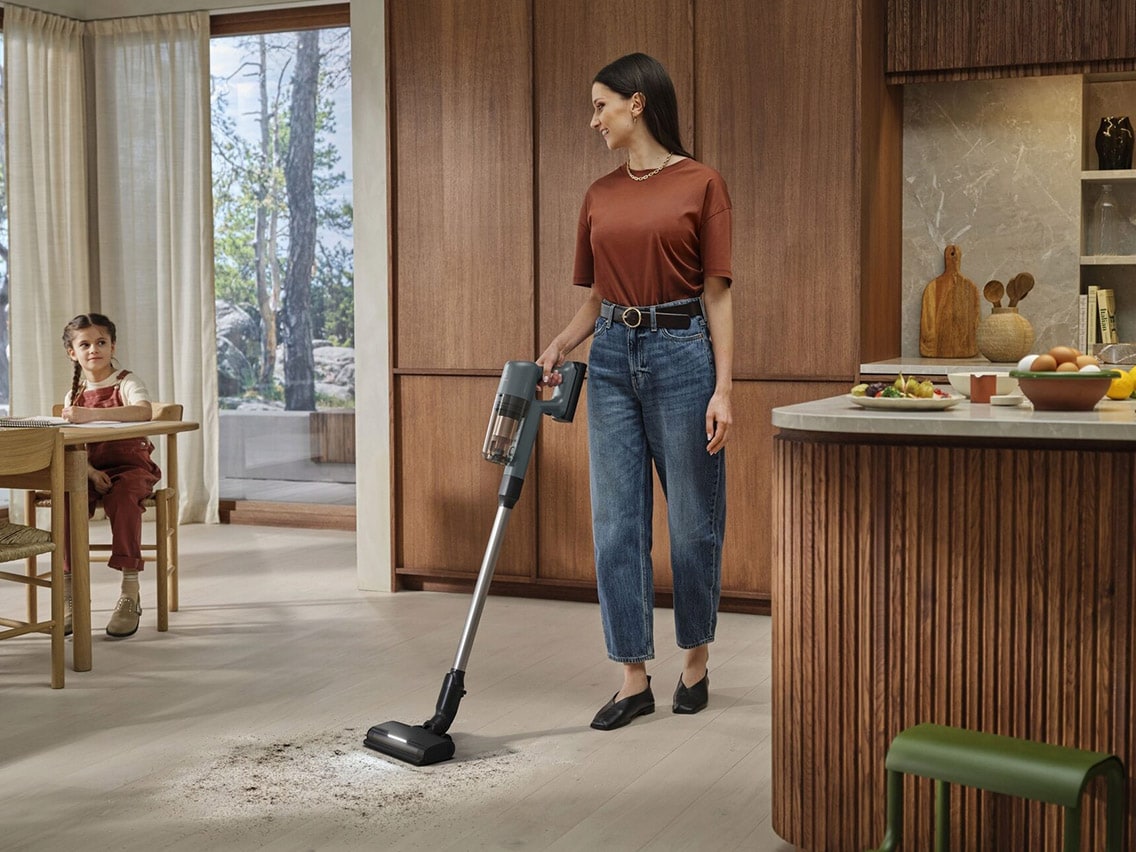 En kvinne bruker en trådløs Electrolux 700-støvsuger til å rengjøre et lyst tregulv i et moderne kjøkken.